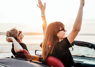 Couple in a convertible with lady raising arms in the air with the ocean in background